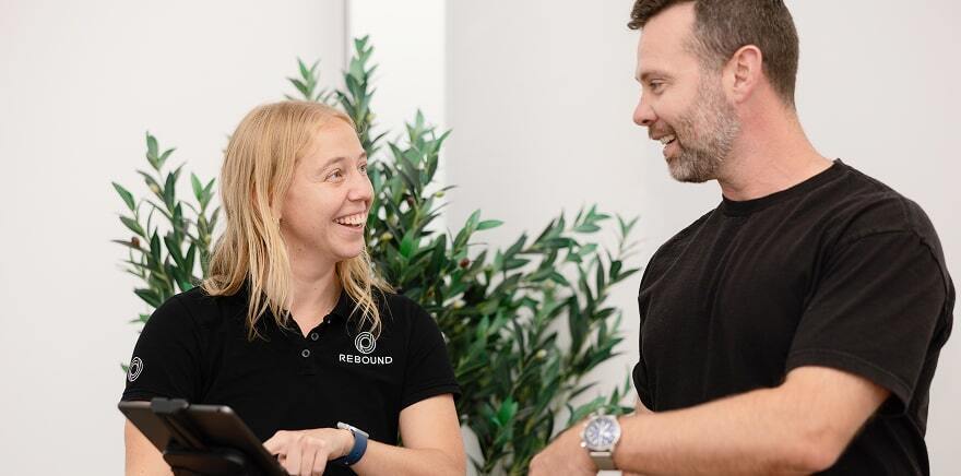 Two people talking indoors, one holding a tablet, with a plant in the background.